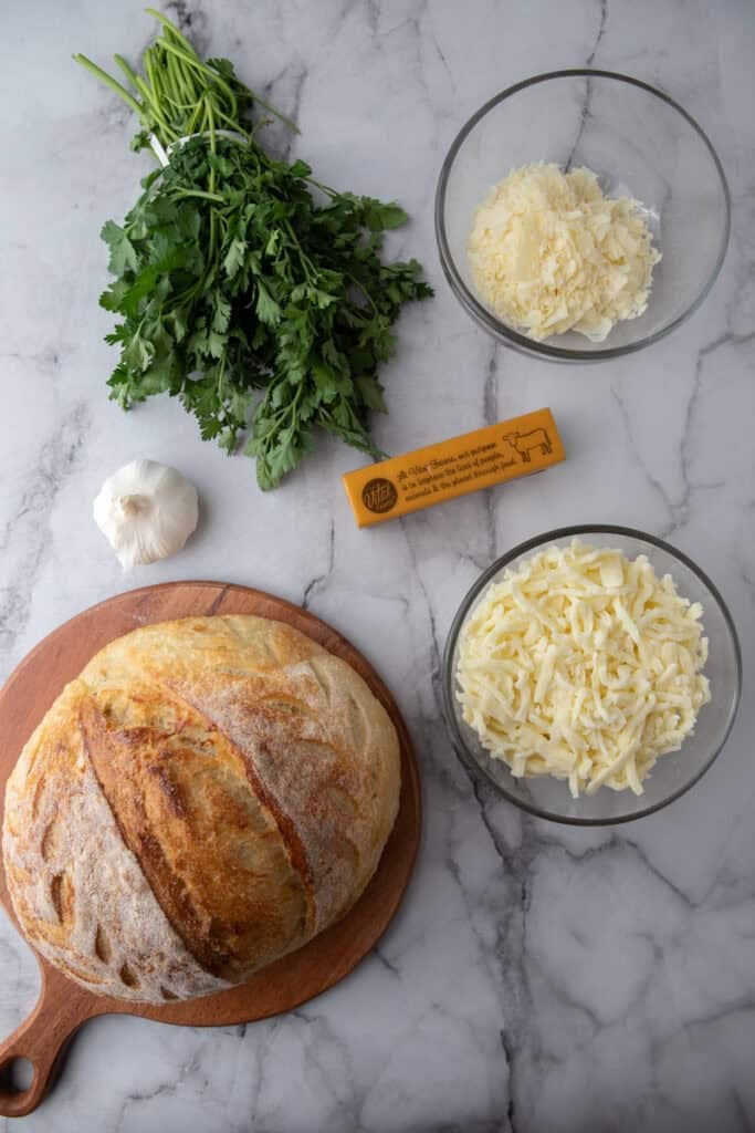 Ingredients for garlic bread including sourdough loaf, mozzarella, parmesan, garlic, butter, and parsley
