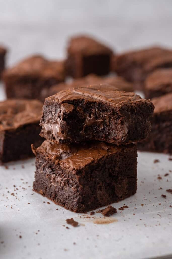 Two fudgy brown butter brownies stacked on parchment paper with melted chocolate chunks visible inside and more brownies in the background