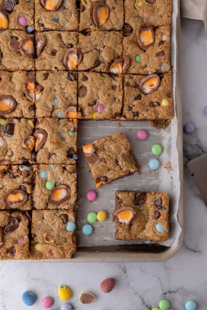 Cadbury egg cookie bars sliced into squares in a parchment lined baking pan with pastel M&Ms and Cadbury creme eggs baked in