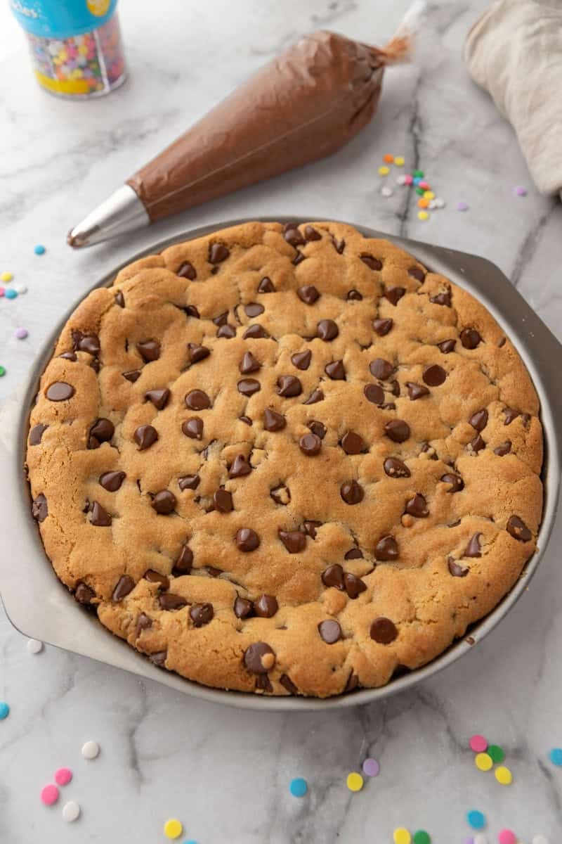 Baked chocolate chip cookie cake in a round pan with a piping bag of chocolate frosting.