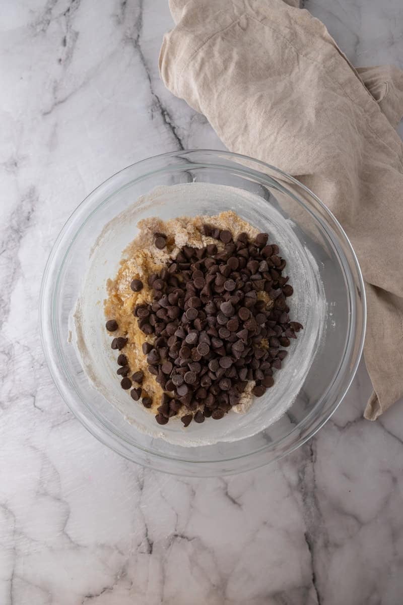 Chocolate chips added to the cookie cake dough in a glass bowl.