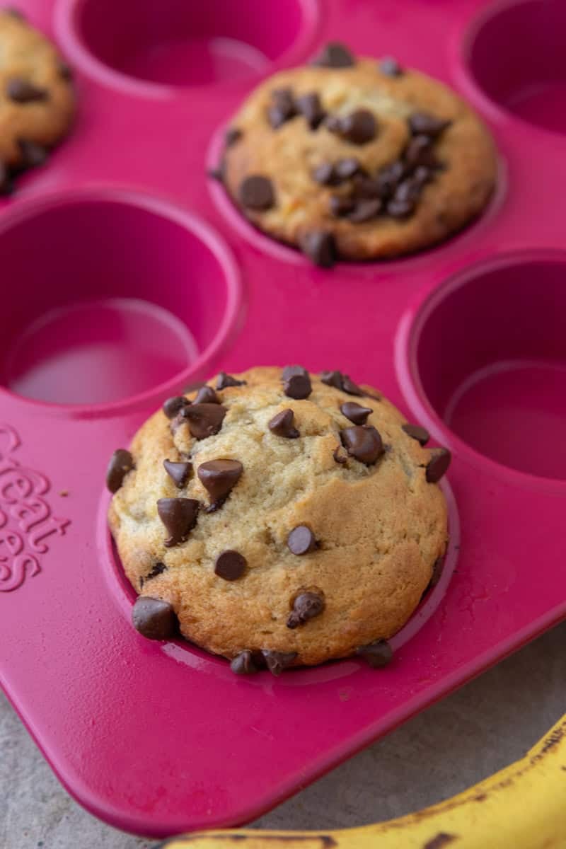 Two baked banana chocolate chip muffins with tall domes in a pink silicone muffin pan.