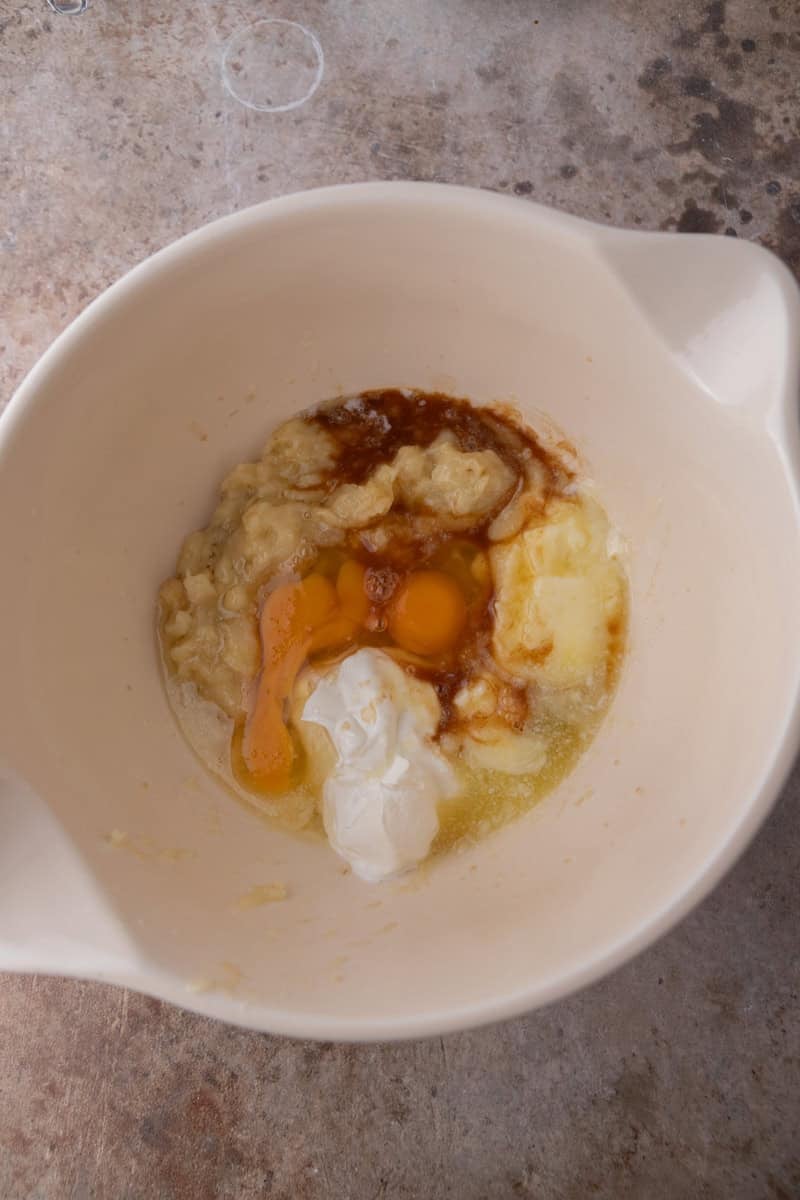 Mashed banana, eggs, melted butter, vanilla, and sour cream in a white mixing bowl.