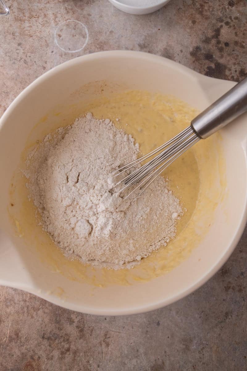 Flour mixture added to the banana batter in a white bowl with a whisk.