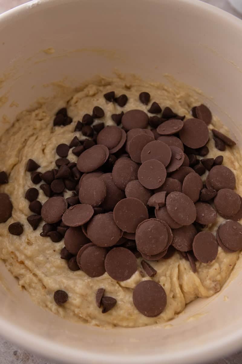Chocolate chips and chocolate discs piled on top of banana muffin batter in a bowl.