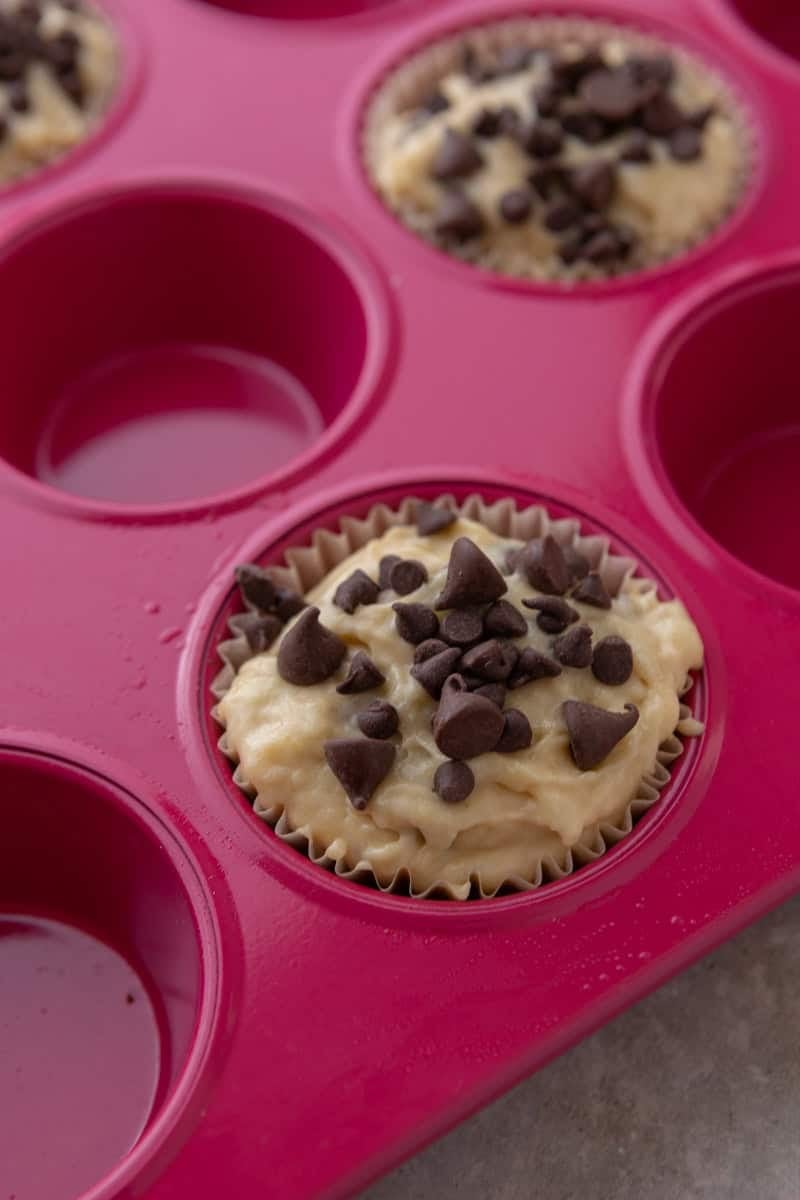 Muffin liners filled to the top with batter and sprinkled with chocolate chips in a pink silicone muffin pan.