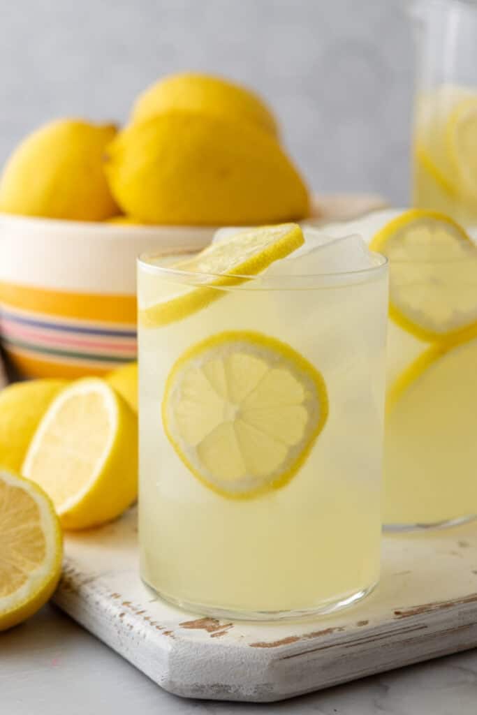 Glass of homemade lemonade with lemon slices and ice, with fresh lemons and a pitcher in the background.