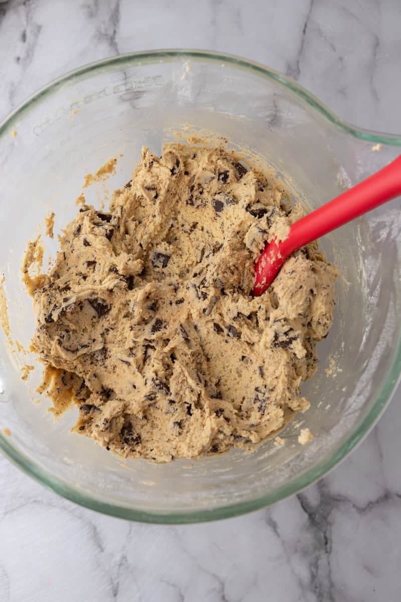 A red spatula folding dark chocolate chunks into the cookie dough.