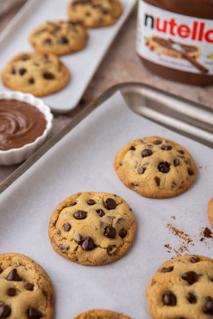 Freshly baked Nutella-stuffed chocolate chip cookies cooling on a tray with Nutella jar in the background
