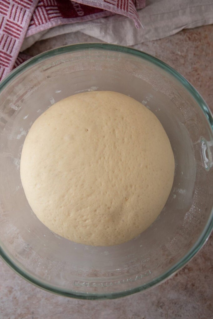 Dinner roll dough doubled in size in a glass bowl after its first rise.