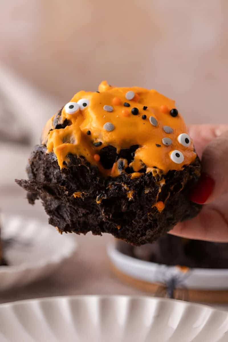 A hand holding a bitten black cinnamon roll with orange frosting, candy eyes, and sprinkles.