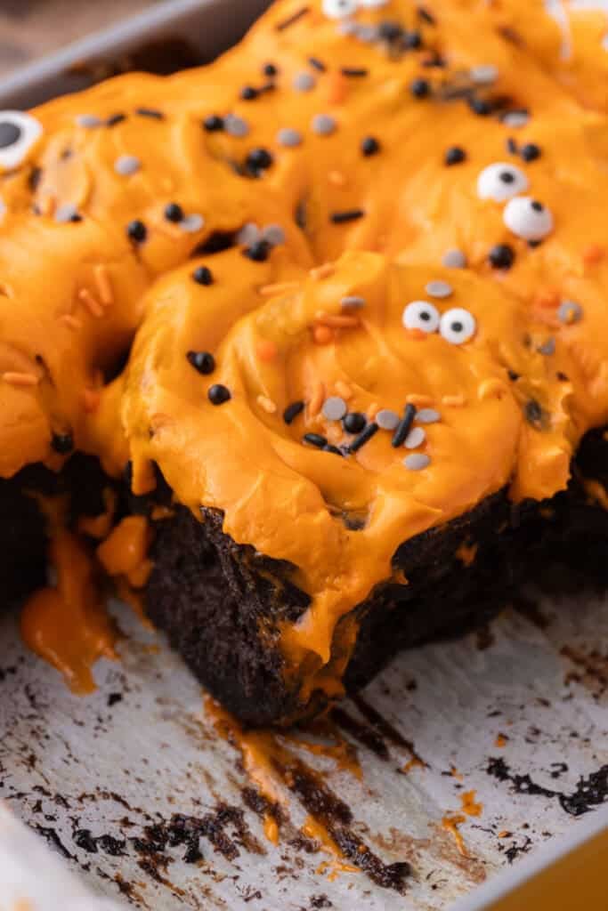 Baked black cinnamon rolls in a pan topped with orange frosting, sprinkles, and candy eyes.
