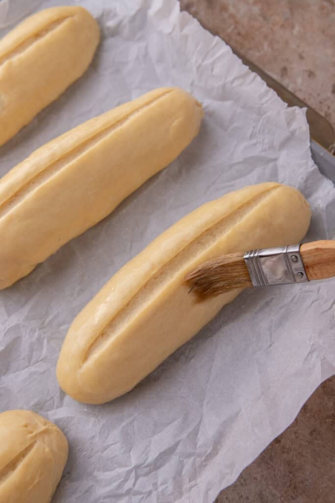Six shaped hoagie rolls doubled in size on a parchment-lined baking sheet with a roll being brushed.