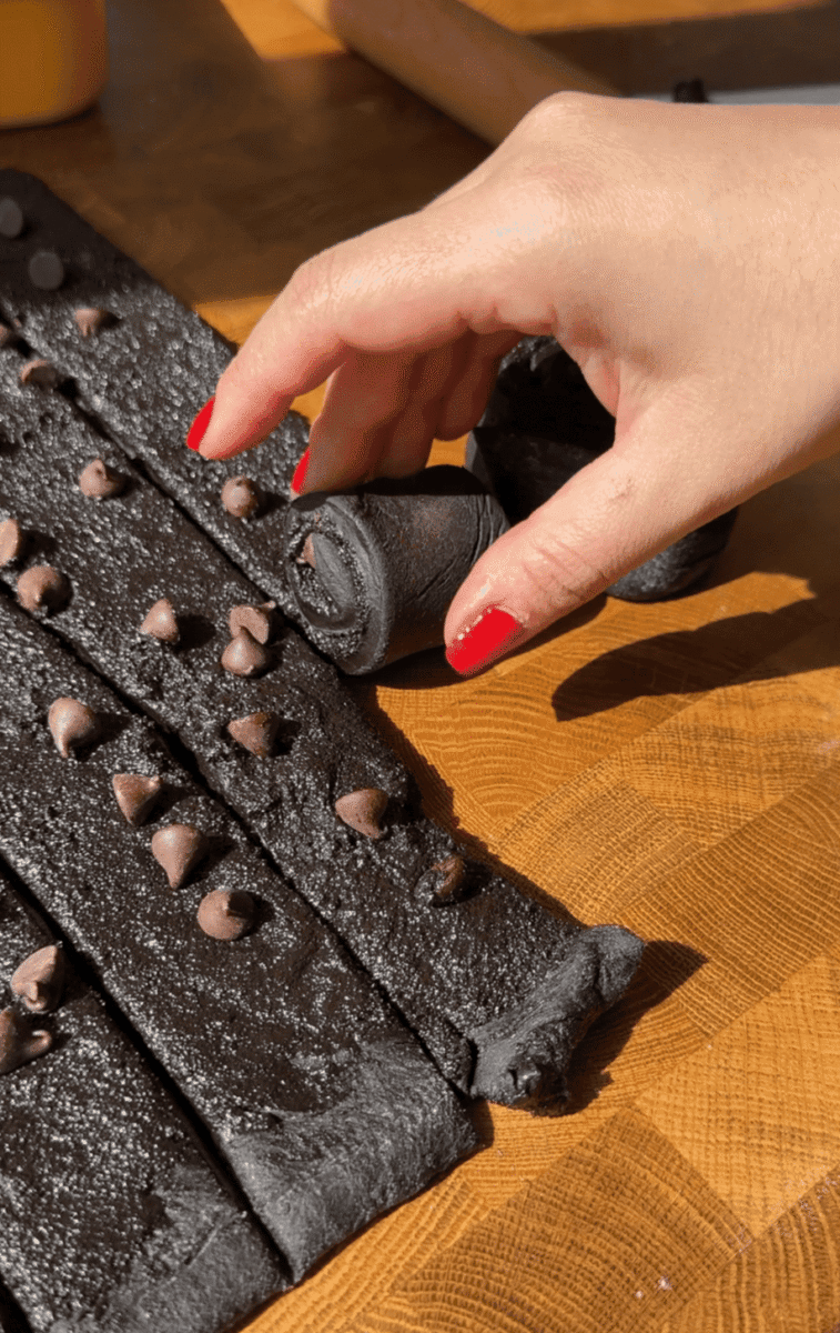 A hand rolling a strip of black cocoa dough filled with cinnamon sugar and chocolate chips.
