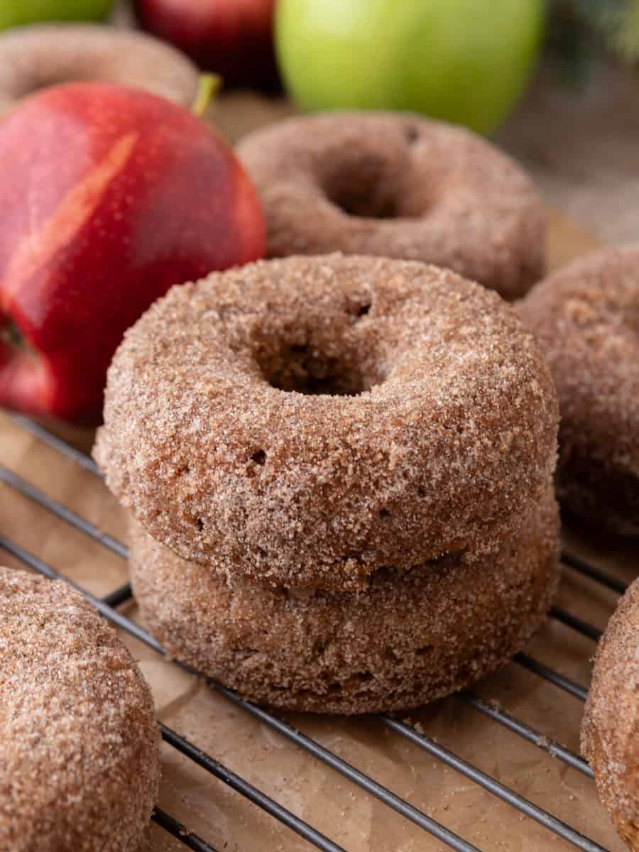 Cinnamon sugar-coated apple cider donuts stacked on a cooling rack with fresh apples in background.