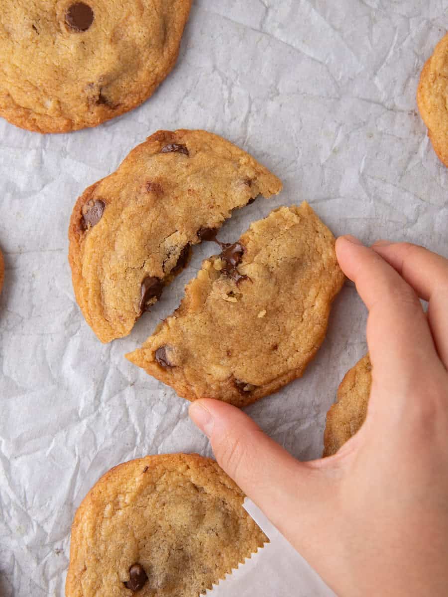 Hand breaking a freshly baked thin chocolate chip cookie showing chewy center.