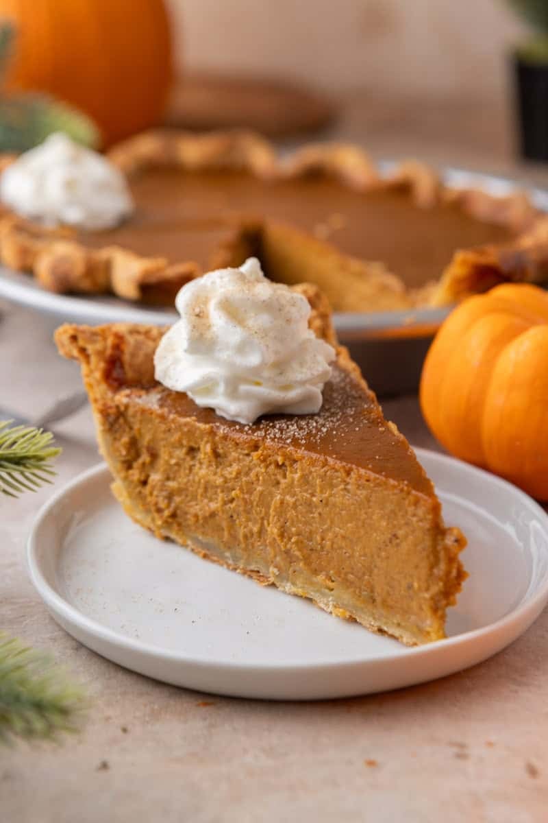 A thick slice of homemade pumpkin pie topped with a swirl of whipped cream and dusted with cinnamon on a white plate.