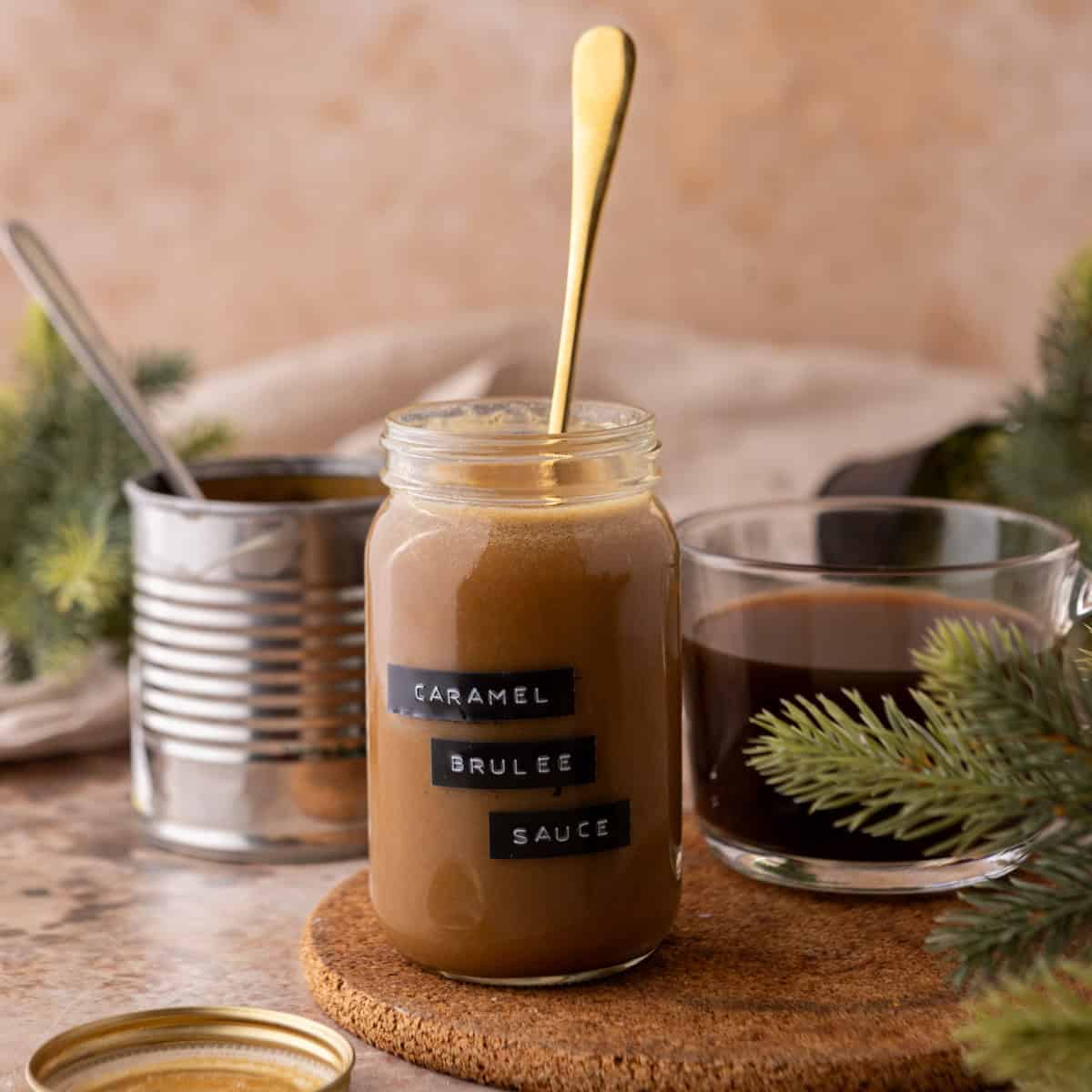 Jar of homemade Starbucks Caramel Brulee Sauce with gold spoon, coffee cup, and festive greenery on a cozy kitchen counter.