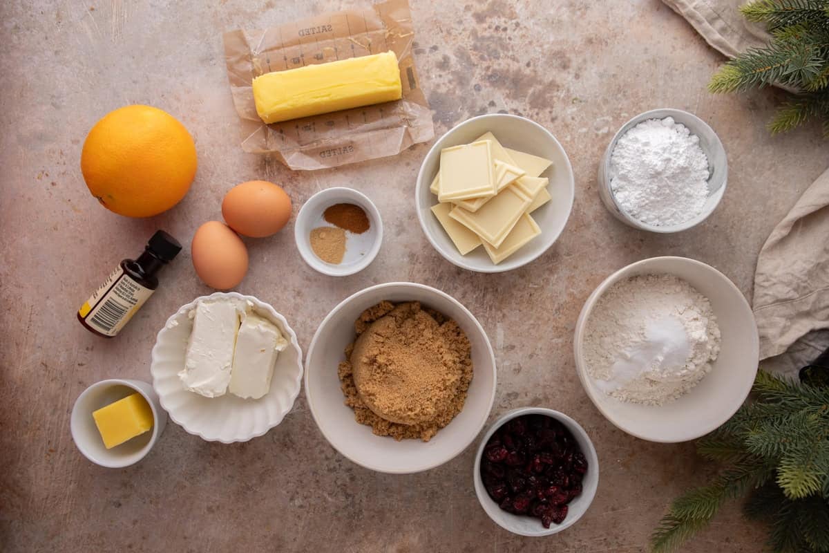 Overhead view of ingredients for cranberry bliss bars including butter, eggs, flour, brown sugar, dried cranberries, white chocolate, orange, spices, cream cheese, and powdered sugar arranged on a counter.