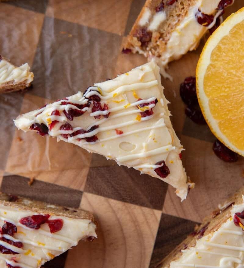 Close-up of a frosted cranberry bliss bar triangle with dried cranberries and orange zest beside a sliced orange.