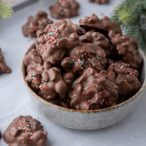 Bowl of chocolate crockpot candy peanut clusters topped with Christmas sprinkles.