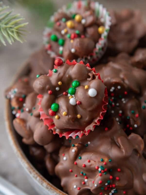Chocolate crockpot candy peanut clusters in mini holiday cupcake liners topped with colorful sprinkles.