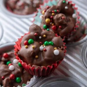 Chocolate crockpot candy peanut clusters in mini holiday cupcake liners topped with colorful sprinkles.