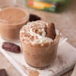 Date Sweetened Hot Chocolate topped with whipped cream and chocolate dates sitting in a short glass on a white wooden board.