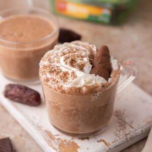 Date Sweetened Hot Chocolate topped with whipped cream and chocolate dates sitting in a short glass on a white wooden board.