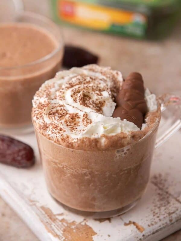 Date Sweetened Hot Chocolate topped with whipped cream and chocolate dates sitting in a short glass on a white wooden board.