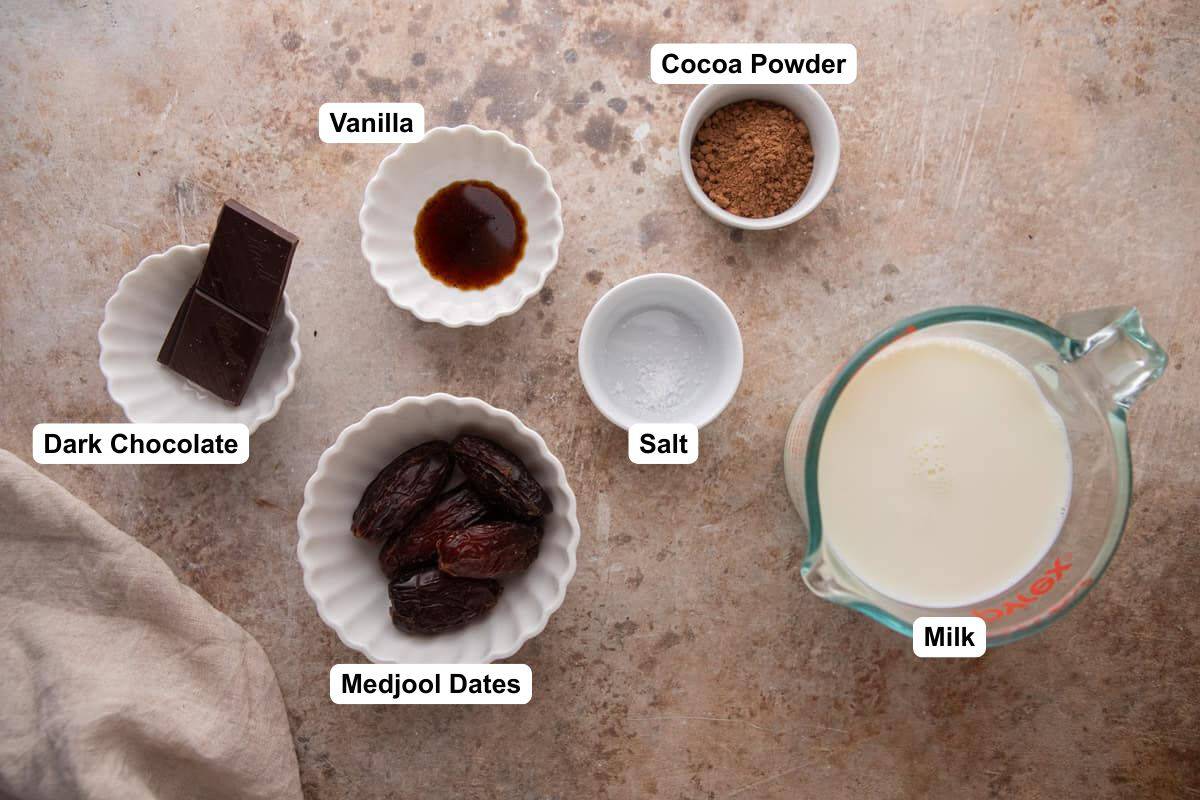 Medjool dates, cocoa powder, dark chocolate, milk, vanilla, salt and whipped cream arranged on a countertop for date sweetened hot chocolate.