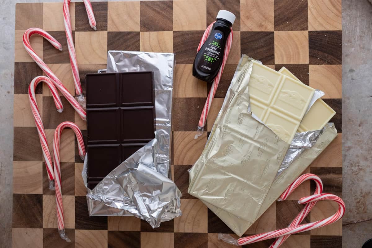 Dark chocolate bars, white chocolate bars, candy canes, and peppermint extract arranged on a wooden board.