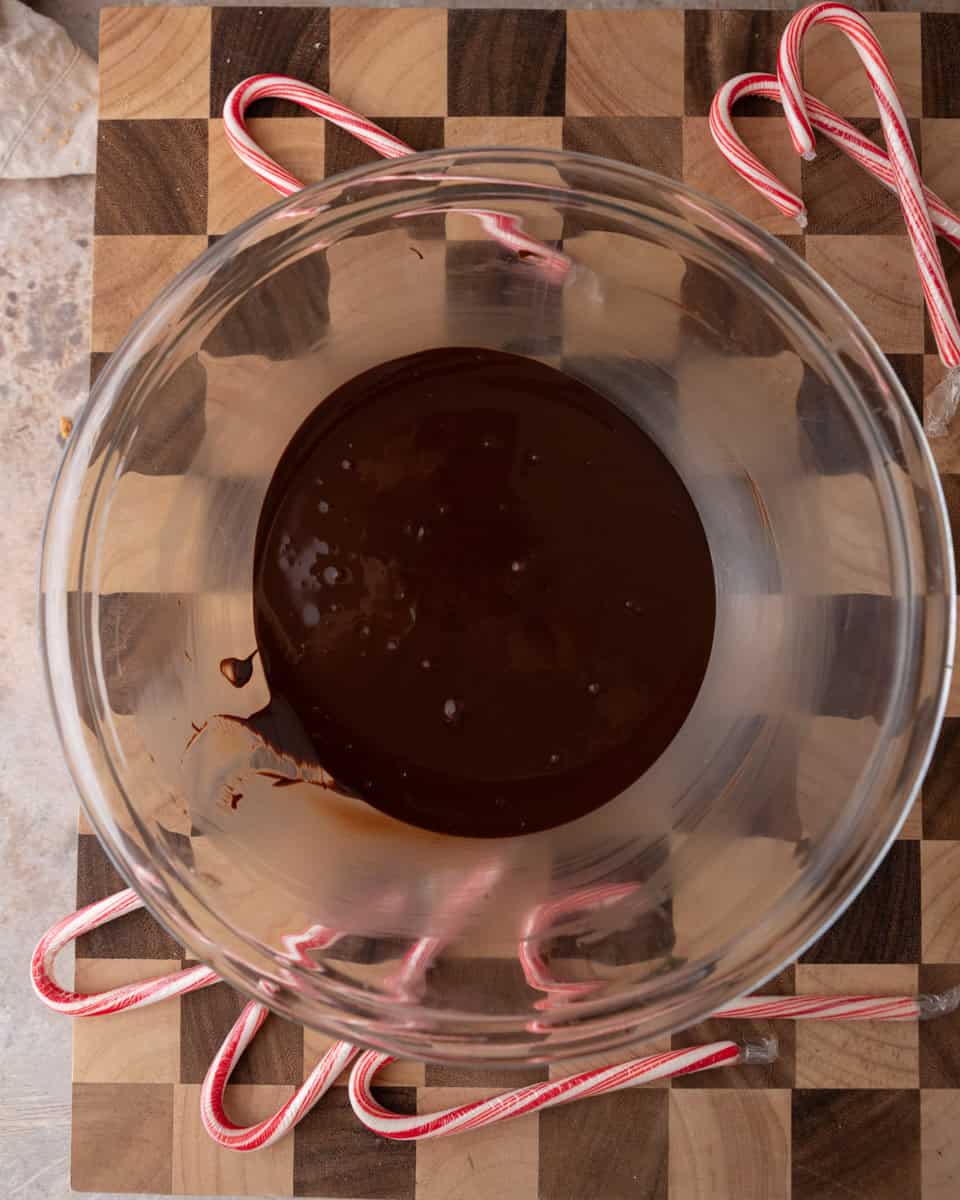 Smooth melted dark chocolate in a glass bowl on a wooden board decorated with candy canes.