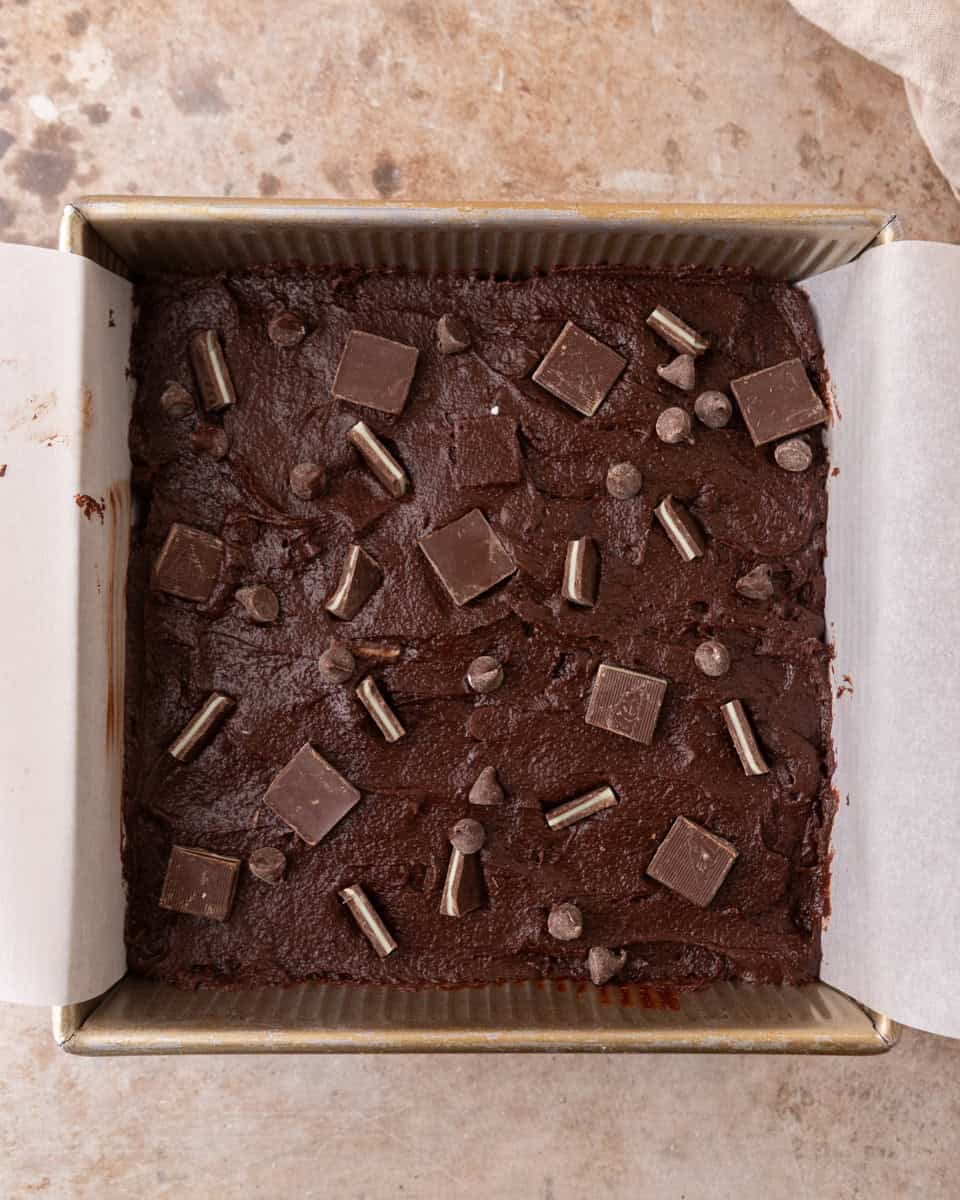  Brownie batter spread in a lined baking pan topped with chocolate and mint pieces