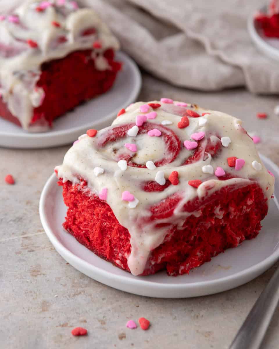 Frosted red velvet cinnamon roll served on a small plate with cream cheese icing and heart sprinkles