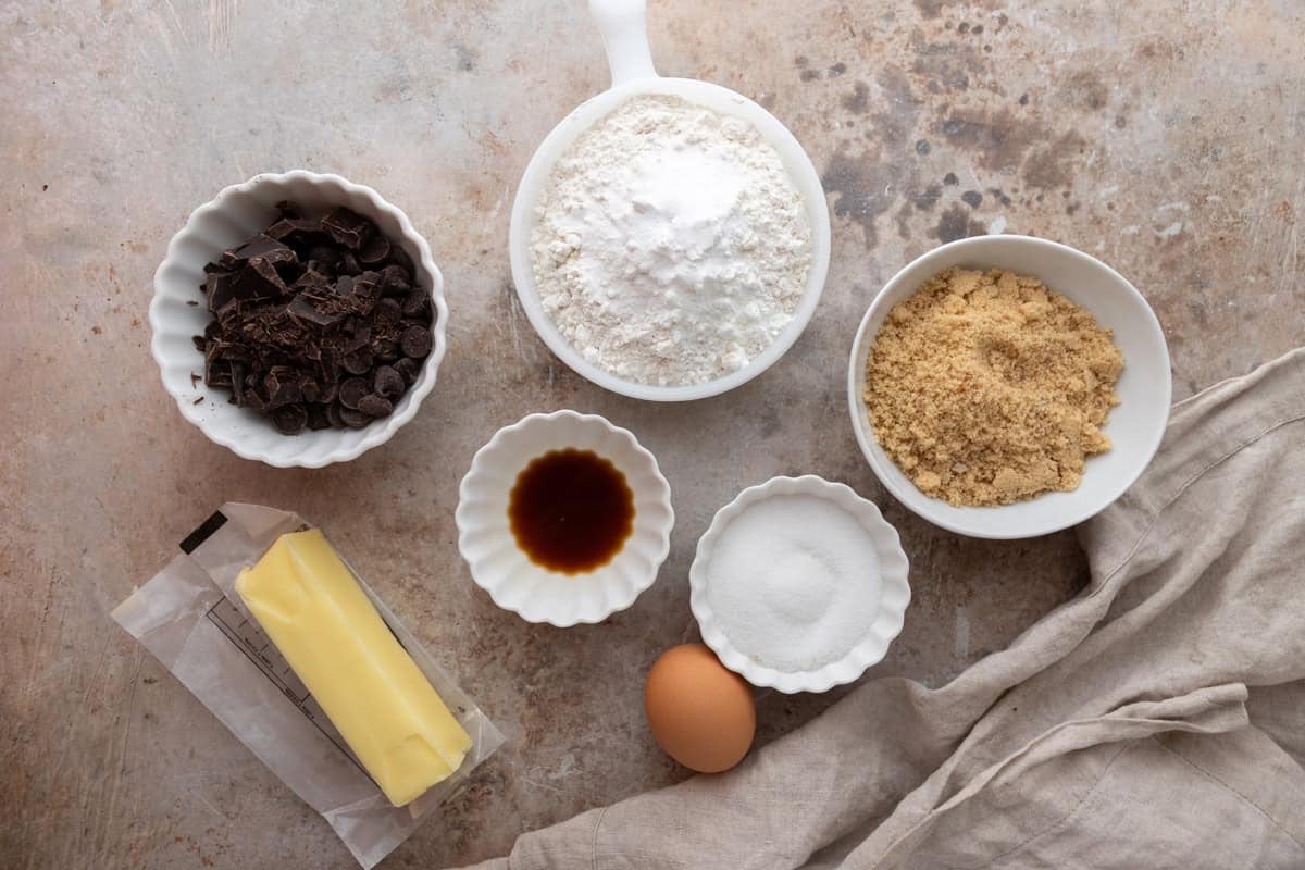 Ingredients laid out on a counter including butter, an egg, chopped chocolate, flour, brown sugar, granulated sugar, and vanilla extract