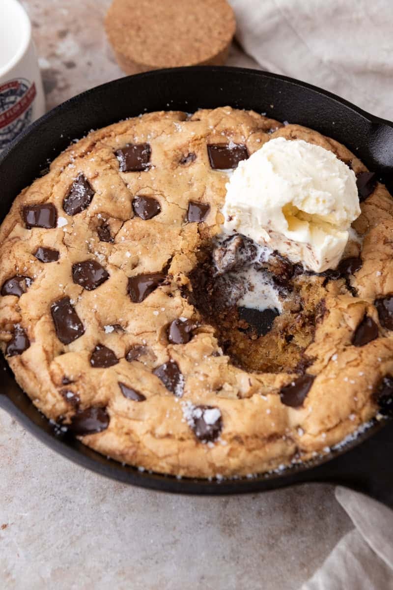 Brown butter chocolate chip skillet cookie in an 8-inch cast iron skillet with a scoop of vanilla ice cream and flaky sea salt with a spoonful scooped out showing the gooey center