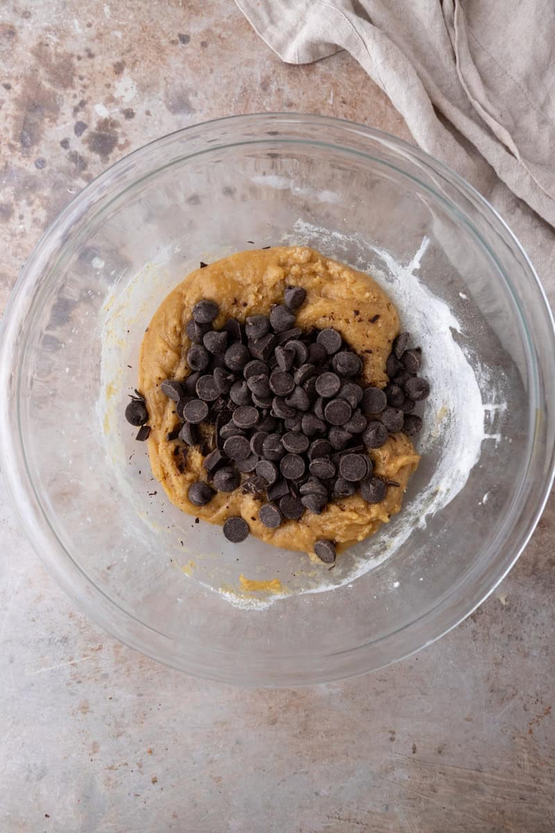 Thick cookie dough in a glass bowl with dark chocolate chips piled on top before folding them in