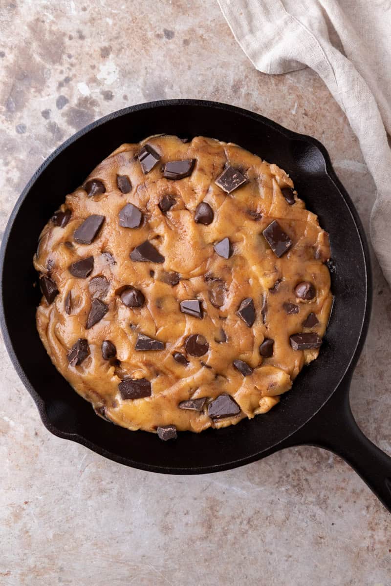 Unbaked chocolate chip cookie dough pressed into an even layer in a greased 8-inch cast iron skillet with chocolate chunks visible on top