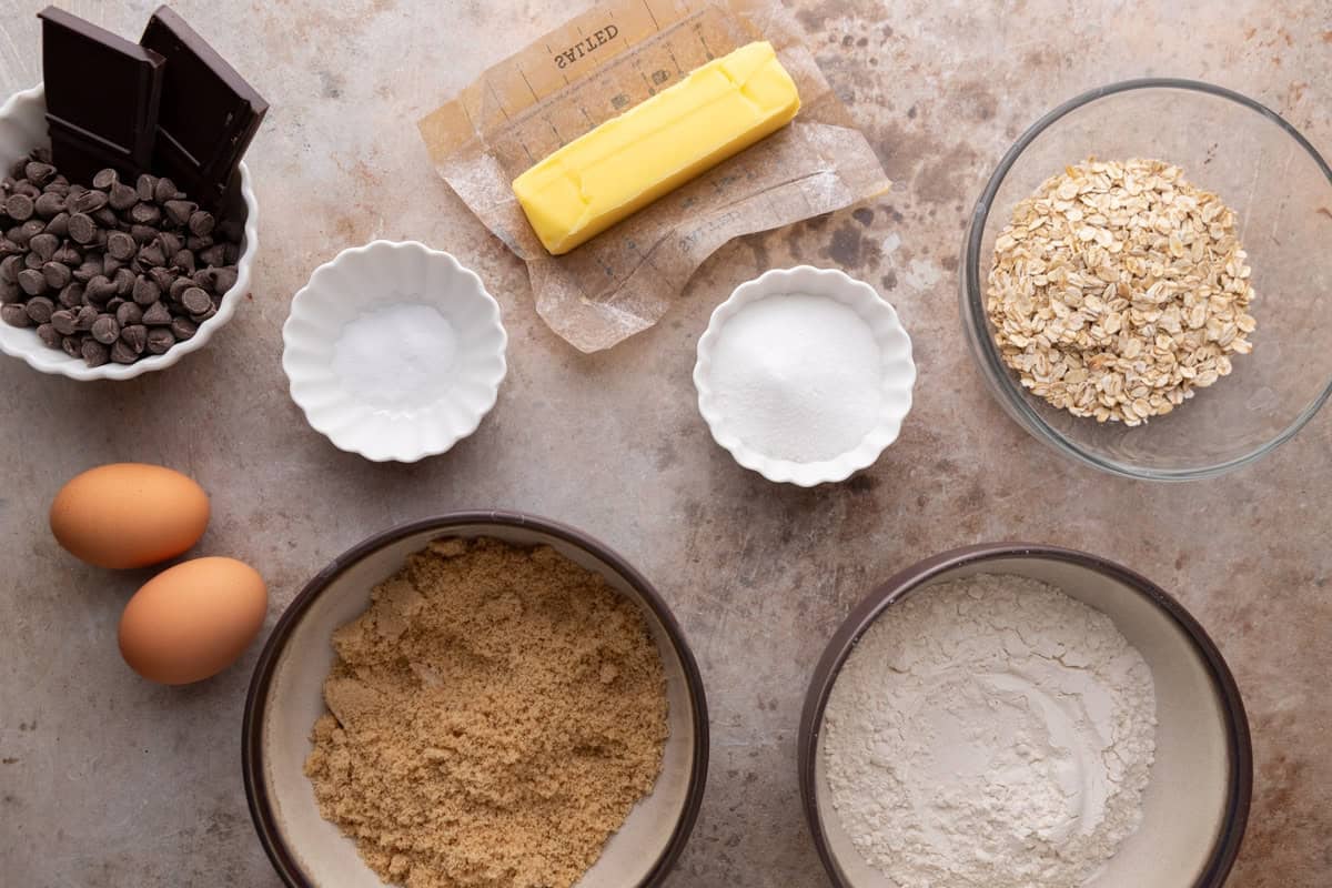 Ingredients for cookie bars laid out in bowls including butter, brown sugar, flour, rolled oats, eggs, chocolate chips, and chocolate chunks