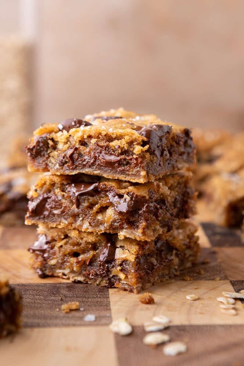 A stack of three thick brown butter oat bars with visible chocolate chunks and scattered oats on a wooden board