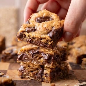 A hand lifting a chewy brown butter cookie bar from a stack on a wooden cutting board, showing melted chocolate and flaky sea salt on top
