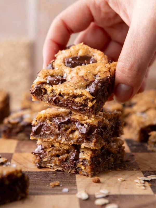 A hand lifting a chewy brown butter cookie bar from a stack on a wooden cutting board, showing melted chocolate and flaky sea salt on top