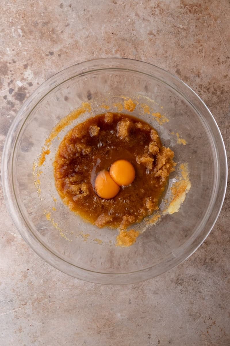An egg and egg yolk added to a whisked brown butter and sugar mixture in a glass bowl