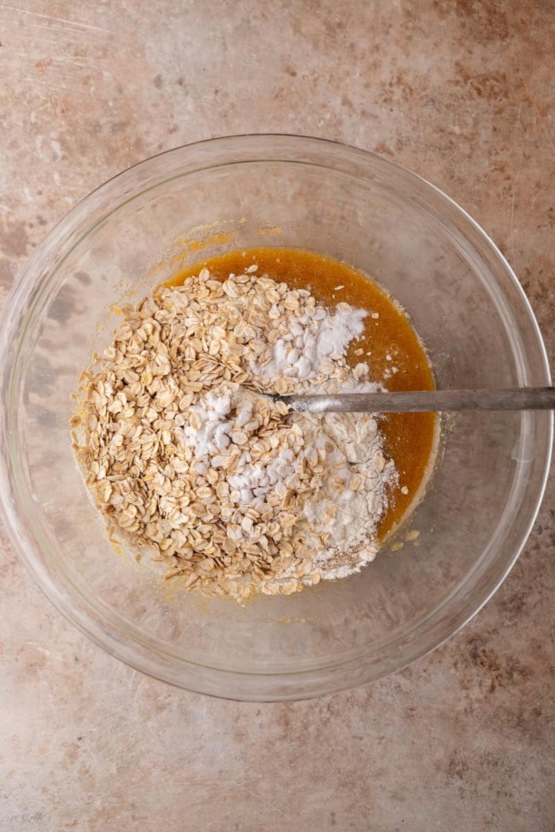 Rolled oats, flour, and baking soda being stirred into wet batter with a spoon in a glass bowl