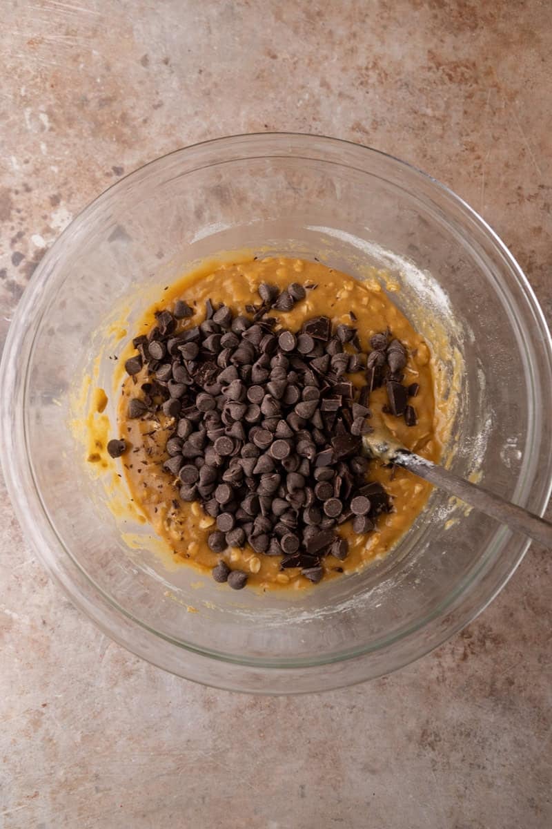 Chocolate chips and chocolate chunks poured on top of cookie bar dough in a glass mixing bowl