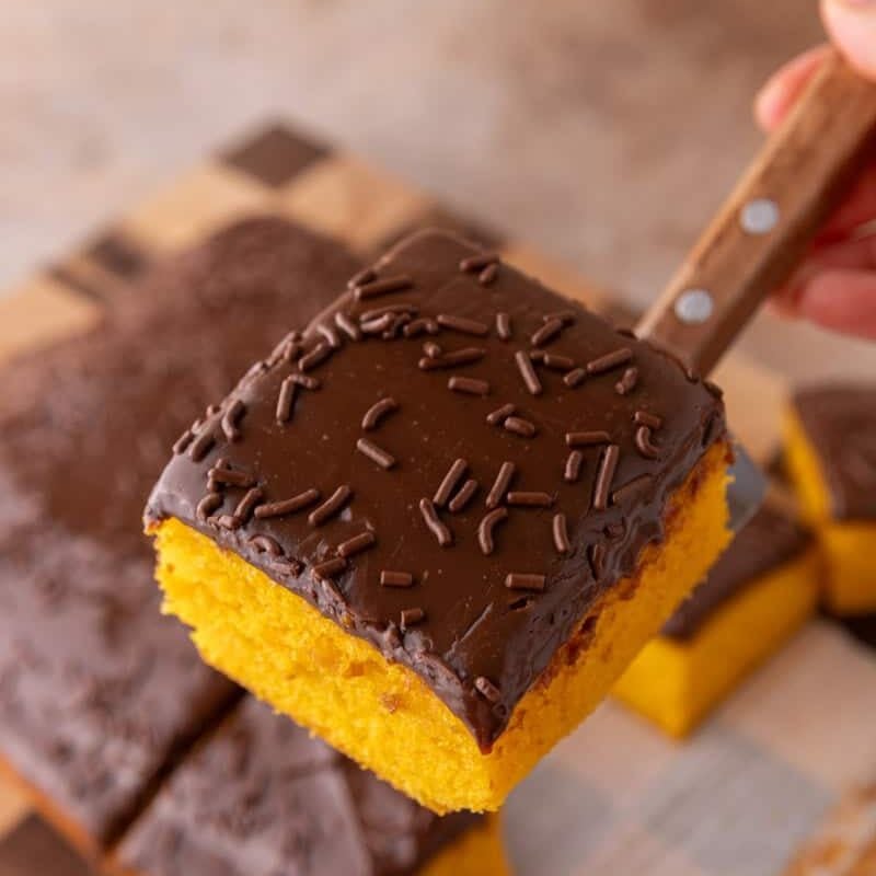 A slice of bright orange carrot cake with thick chocolate glaze and chocolate sprinkles being lifted with a wooden-handled knife
