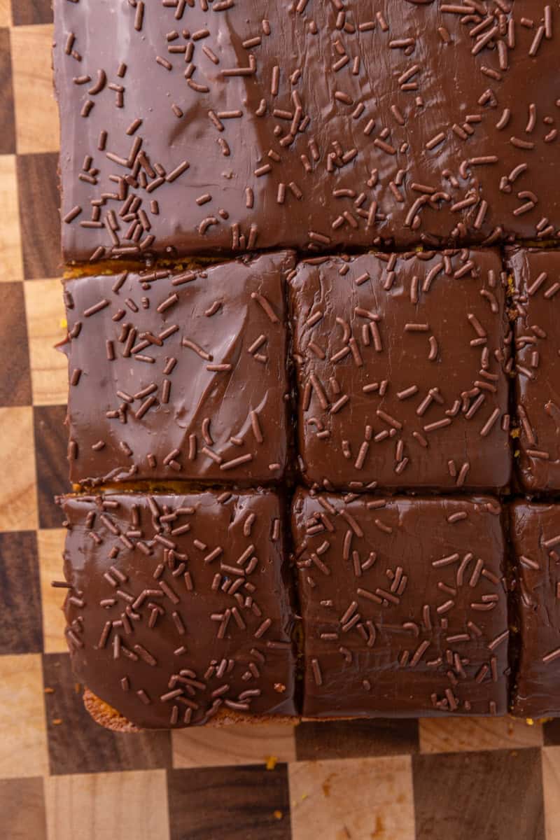 Overhead shot of chocolate-glazed cake sliced into squares on a checkered wood board with chocolate sprinkles on top