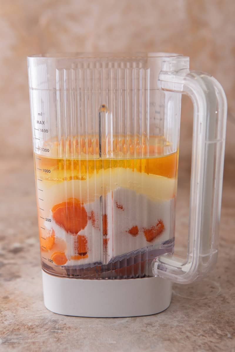 A blender filled with layers of chopped carrots, sugar, oil, and eggs before blending