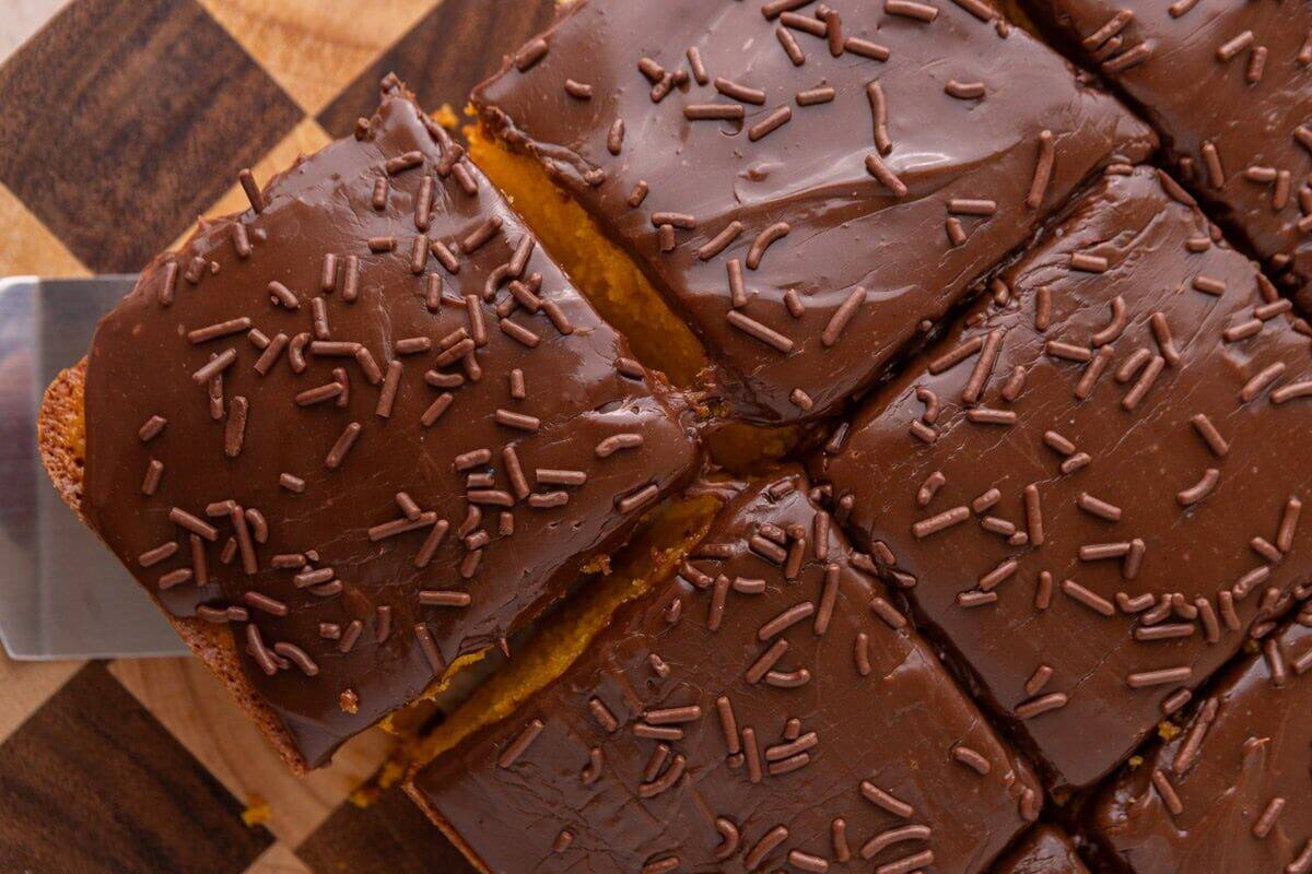 Overhead view of sliced Brazilian carrot cake on a checkered wood cutting board with a spatula lifting one piece